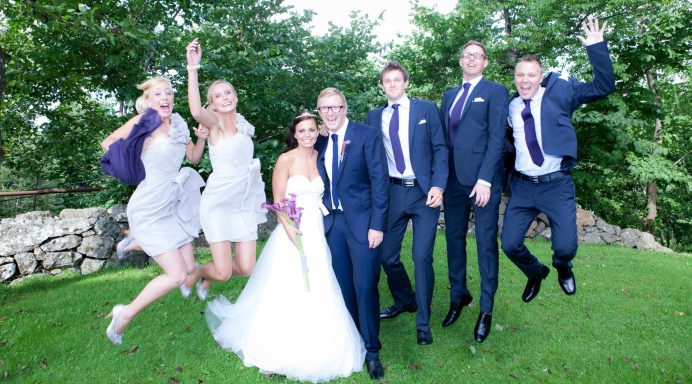 Brudpar och bröllopsfölje hoppar glädjefyllt i en grön trädgård.